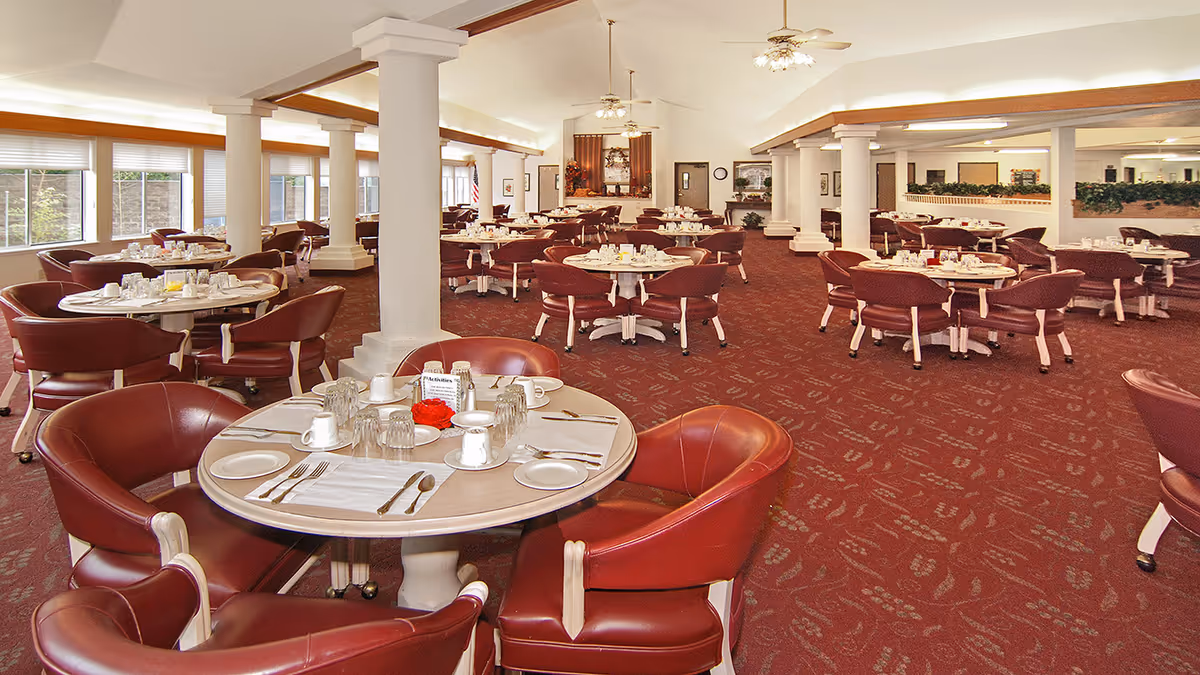 A spacious dining room with multiple round tables set with white plates, cups, and silverware. The room features maroon carpet and matching maroon chairs with white legs. Large windows line the left side of the room, allowing natural light to fill the space. White columns and ceiling fans with lights are visible throughout the room.