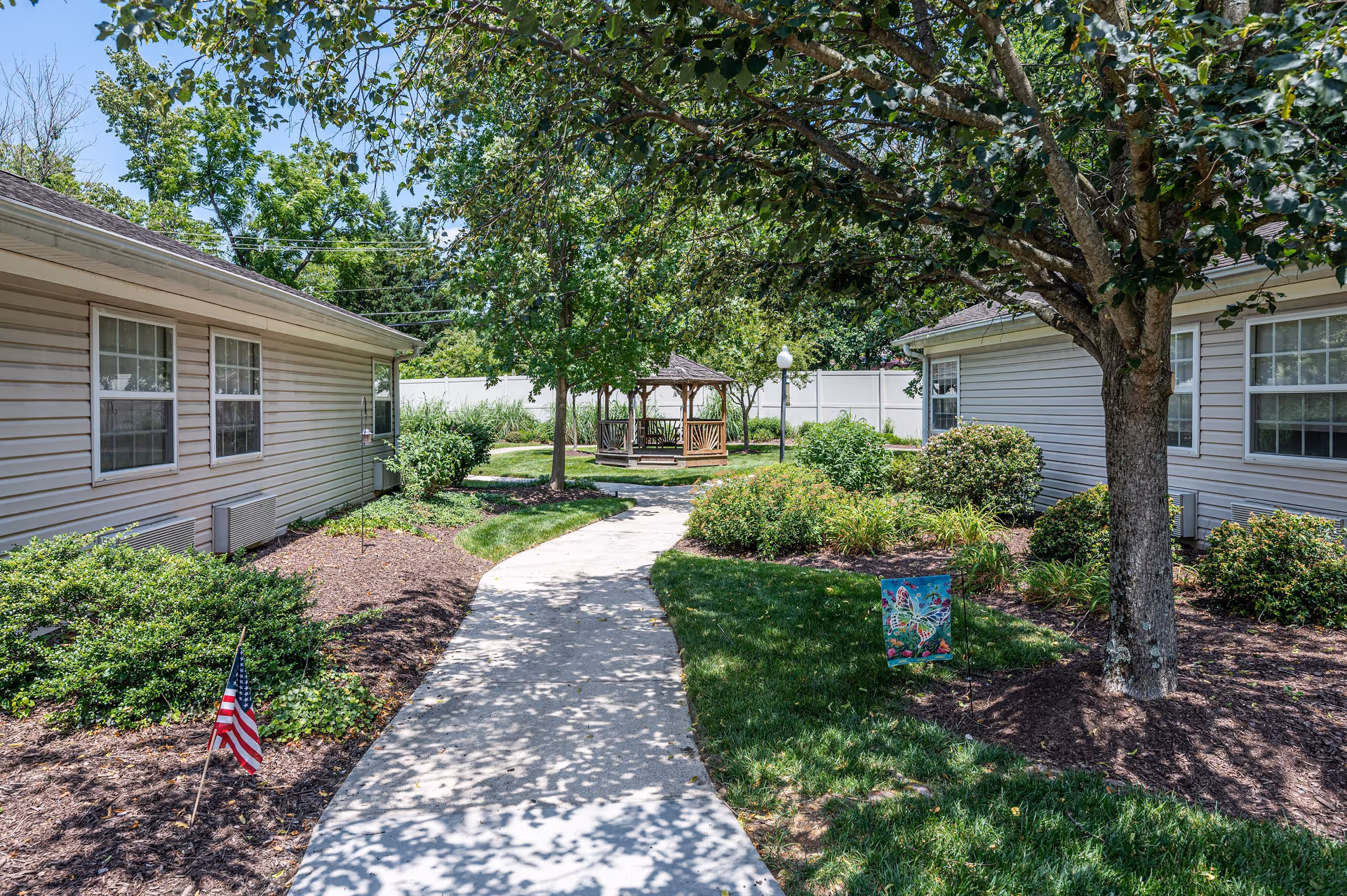 A paved walkway curves through a landscaped garden area between two single-story buildings with beige siding and white-framed windows. The garden features green bushes, a tree casting shadows on the path, and a small American flag planted in the mulch. In the background, there is a wooden gazebo surrounded by more greenery and a white fence enclosing the area.