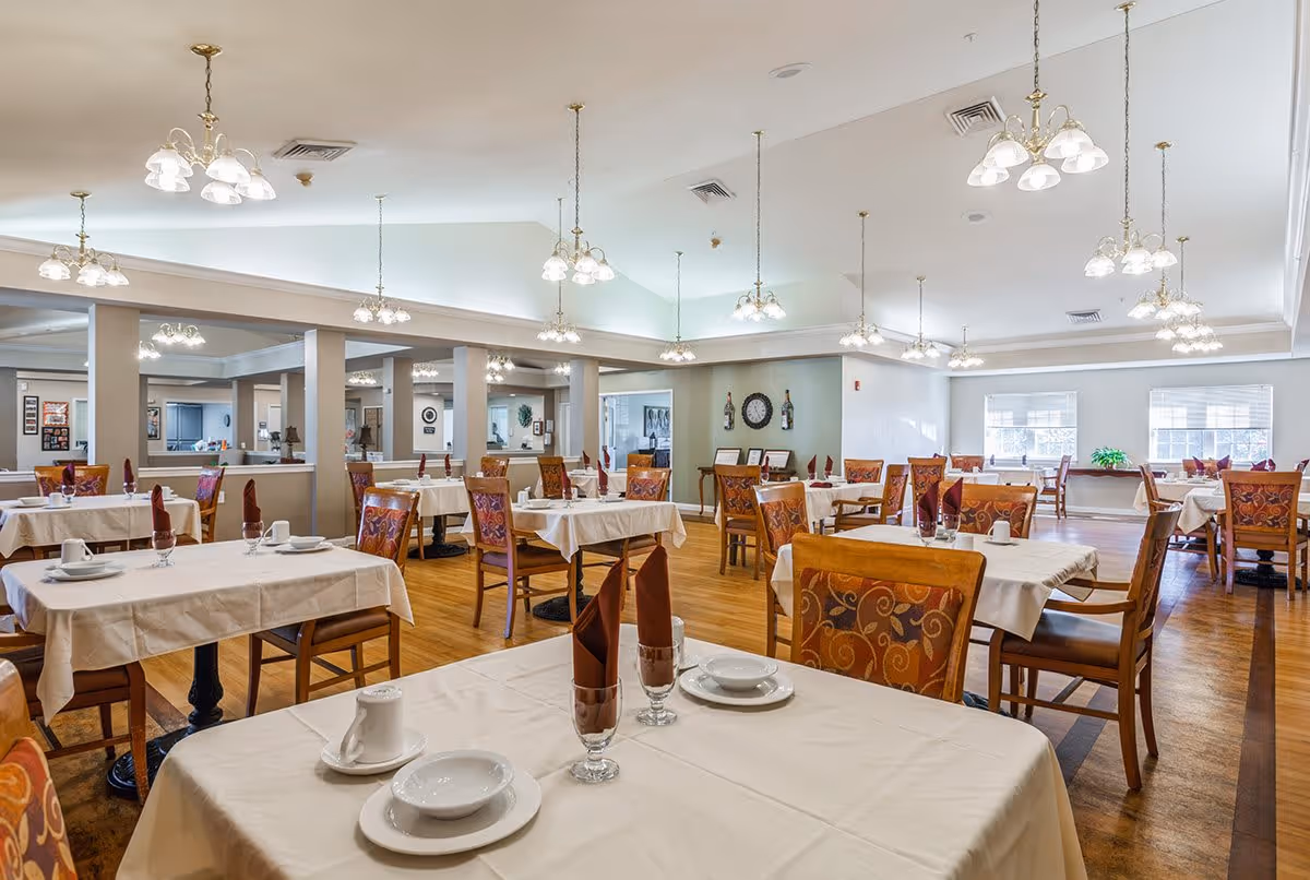 A spacious dining room with multiple tables covered in white tablecloths, each set with plates, bowls, cups, and glasses with folded maroon napkins. The room features wooden chairs with patterned upholstery, hardwood floors, and multiple ceiling light fixtures. Large windows allow natural light to brighten the space.