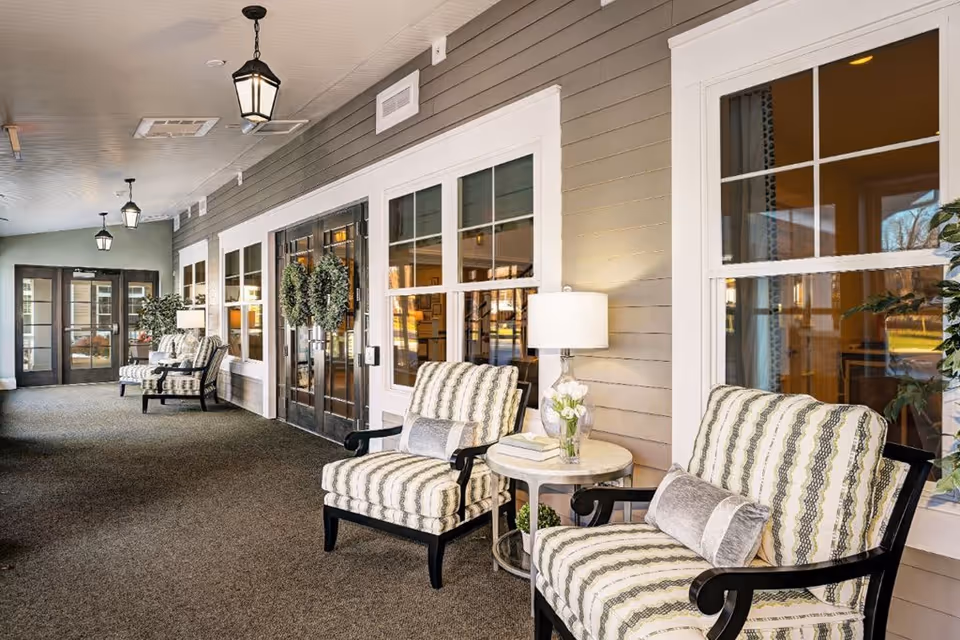 A bright and inviting indoor seating area with patterned armchairs, a small round table with a lamp and flowers, large windows, and double doors decorated with wreaths. The space has a carpeted floor and hanging lantern-style ceiling lights.