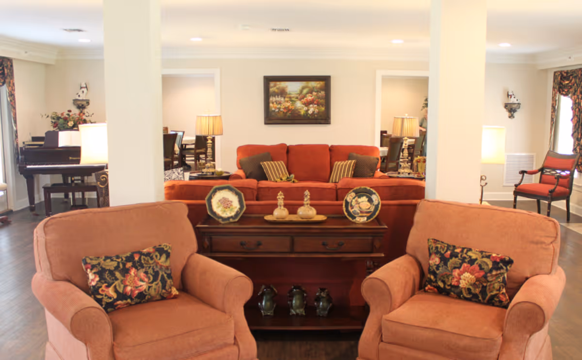 A cozy living room area in an assisted living facility featuring two orange armchairs with floral cushions in the foreground, a wooden console table with decorative plates and figurines, and an orange sofa with striped and solid cushions in the background. The room has hardwood floors, a piano on the left side, lamps, floral curtains, and framed artwork on the wall.