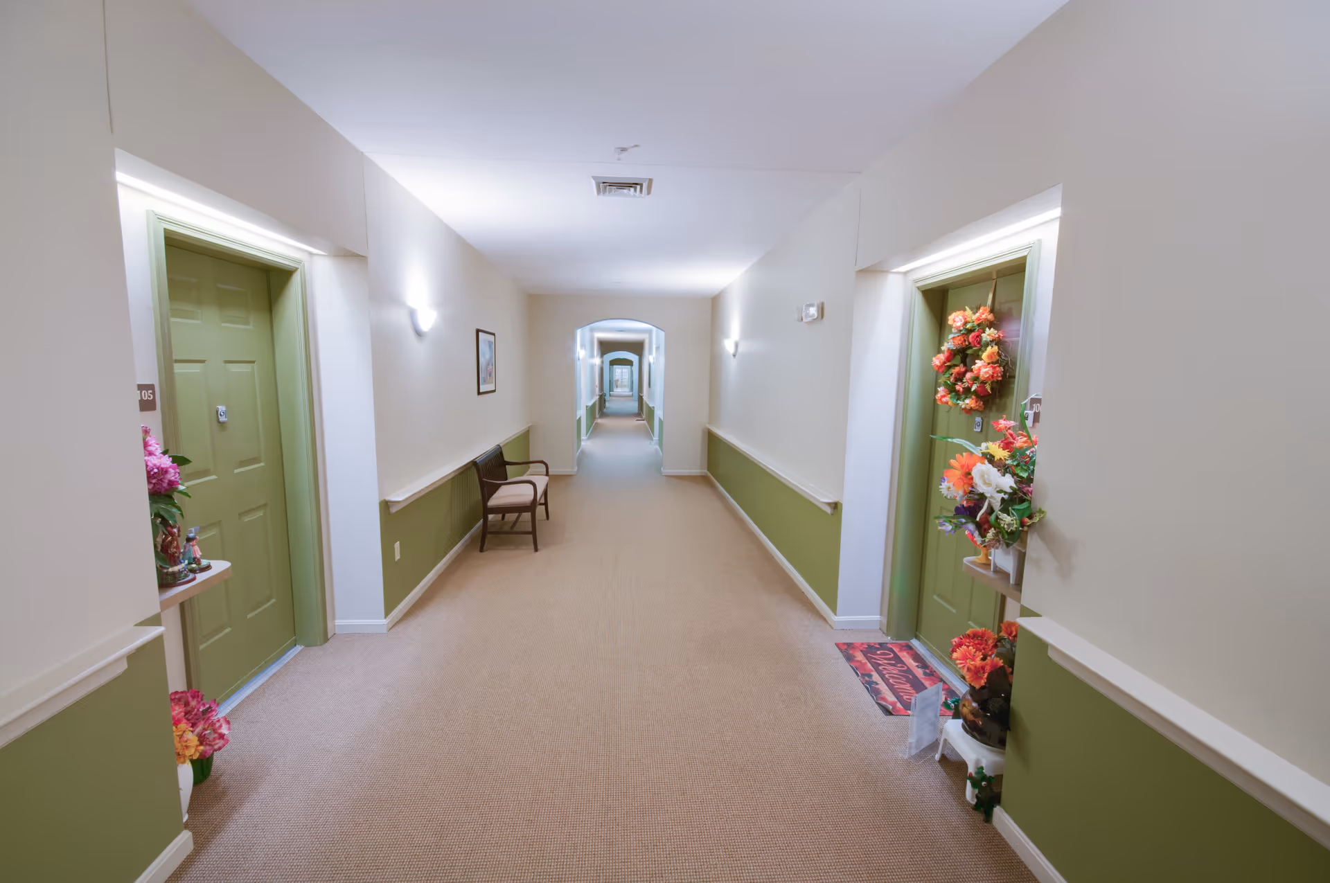 Carpeted interior corridor of a senior living facility with green apartment doors, a chair, and floral door decorations.