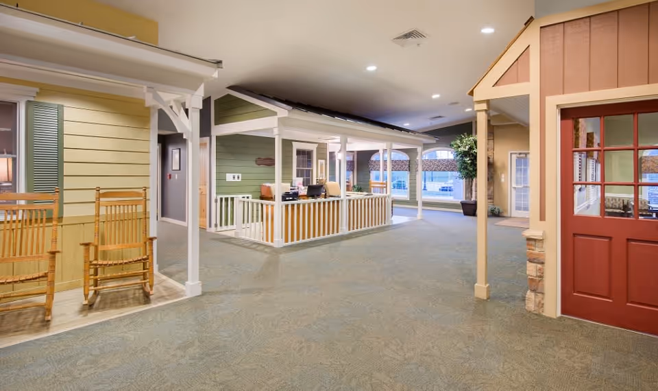 Interior view of a senior living facility designed to resemble a small neighborhood with house-like structures, including a green house with a porch and a red door house. The area is carpeted and well-lit with ceiling lights, and there are two wooden rocking chairs placed near the green house. Large windows and a potted plant are visible in the background.