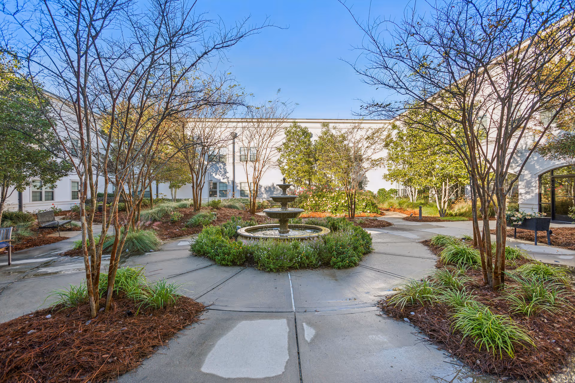 Outdoor courtyard area with a central tiered water fountain surrounded by greenery and trees. Concrete pathways radiate outward from the fountain, with benches and landscaped garden beds along the sides. The courtyard is enclosed by a white building under a clear blue sky.