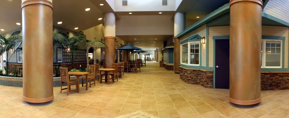 Indoor common area with large columns, tiled floor, wooden tables and chairs, and small house-like structures with windows and doors. The space is well-lit with ceiling lights and has some indoor plants along the sides.