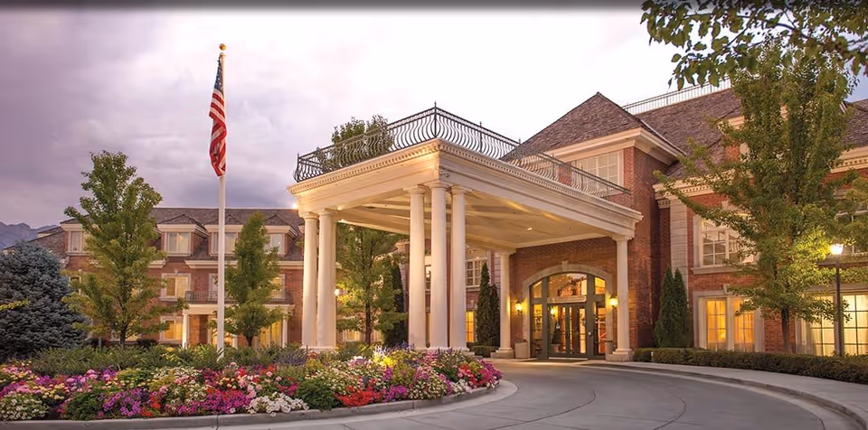Entrance of a brick senior living building with a columned portico, circular driveway, American flag, and manicured flowerbeds.