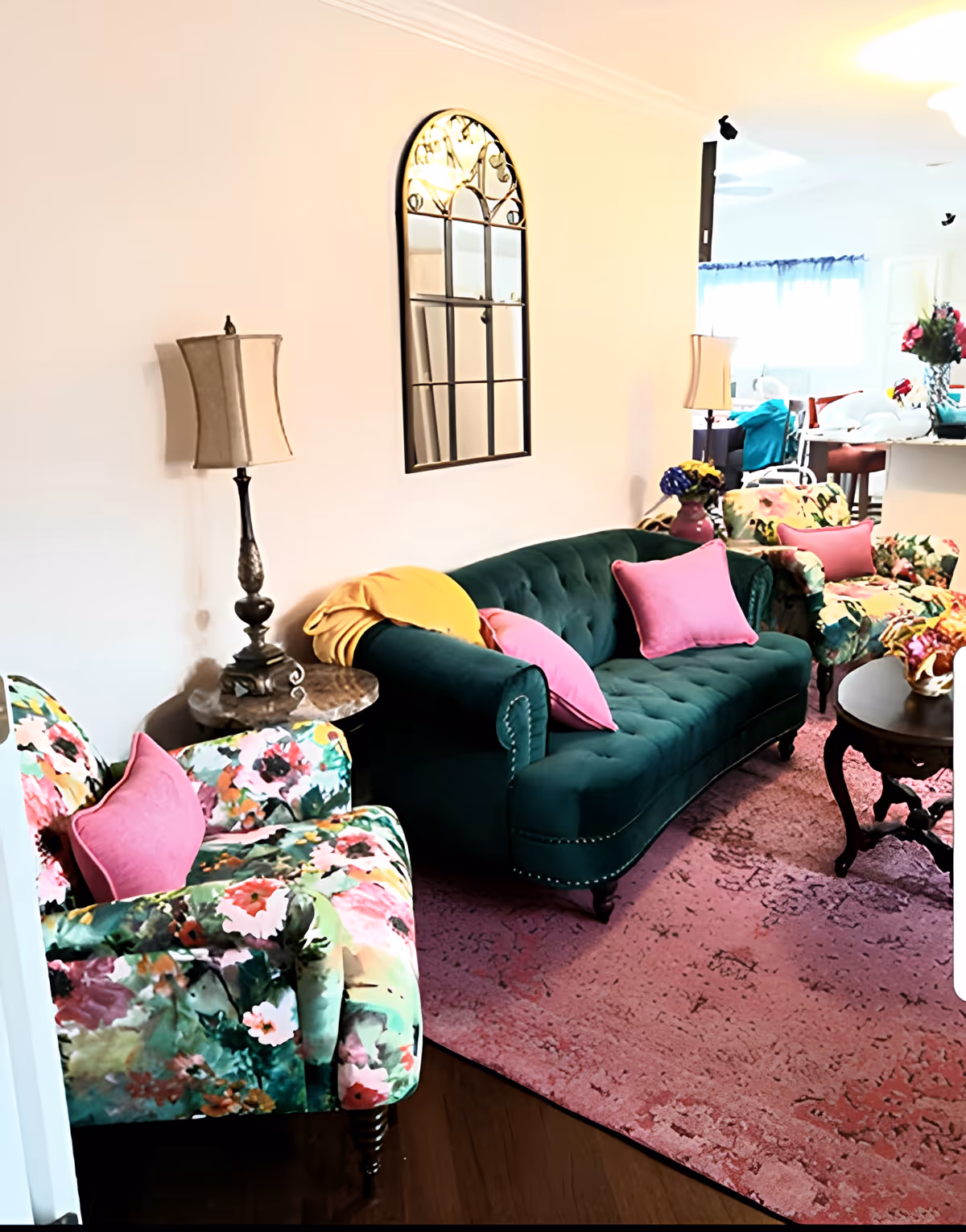 A bright living room with a green tufted sofa, floral armchairs, pink throw pillows, lamps, a decorative wall mirror, and a pink area rug.