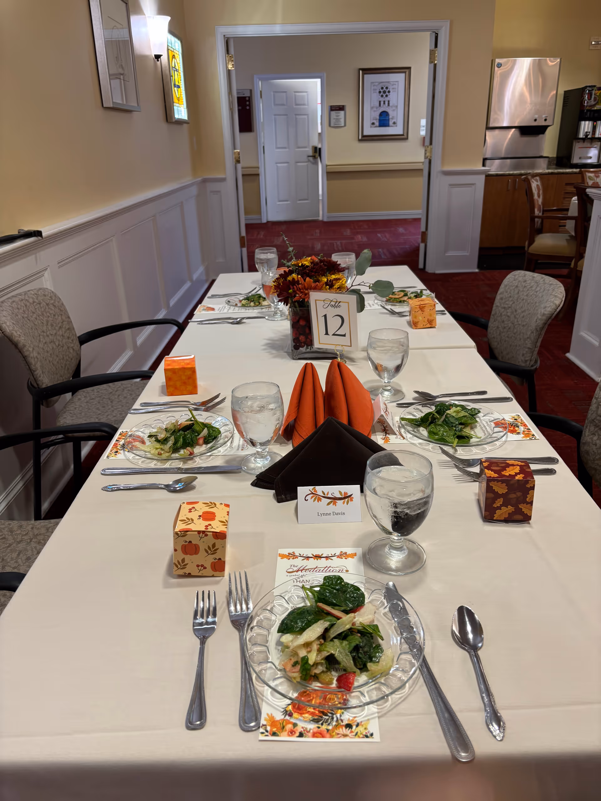 A dining table set for six with white tablecloth, glass plates with salad, water glasses, silverware, and fall-themed decorations including folded orange and brown napkins, small decorative boxes, and a floral centerpiece. The table is in a room with beige walls, carpeted floor, and chairs around the table. A door and framed pictures are visible in the background.
