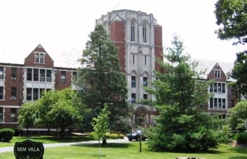 Front view of a multi-story brick building with a central tower, lawn, trees, and a sign reading 'SEM VILLA'.