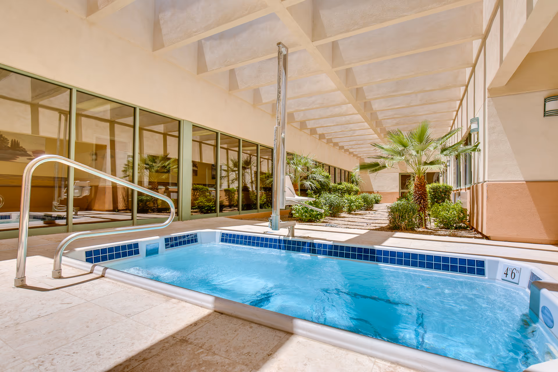 Outdoor hot tub with clear blue water next to a building with large windows and a covered patio area featuring palm trees and shrubs.
