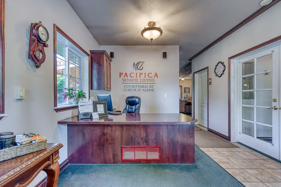 Reception area of Pacifica Senior Living Courtyard at Coeur d'Alene featuring a wooden front desk with a black office chair behind it, a wall clock on the left wall, a window with a plant on the windowsill, and a sign on the back wall displaying the facility name. The area has carpeted and tiled flooring, with a ceiling light fixture and a glass door on the right.