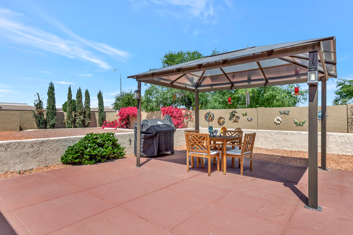 Outdoor patio area with a transparent roof pergola covering a wooden table and four chairs. There is a covered barbecue grill next to the table. The patio is surrounded by a low beige wall decorated with colorful butterfly and flower wall art. Green bushes and pink flowering plants are visible along the wall, with tall trees and a clear blue sky in the background.
