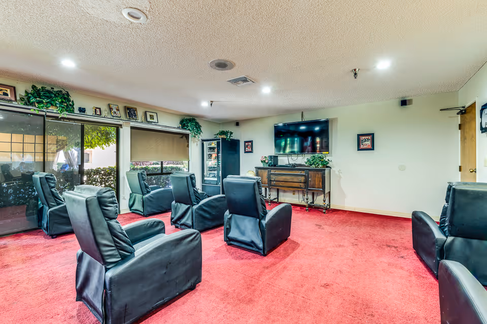 A common area in Santa Anita Assisted Living with six black leather recliners arranged facing a wall-mounted flat screen TV. The room has red carpet flooring, light-colored walls, and several framed pictures and plants placed on a wooden console and shelves. Large windows with blinds and sliding glass doors allow natural light into the space.