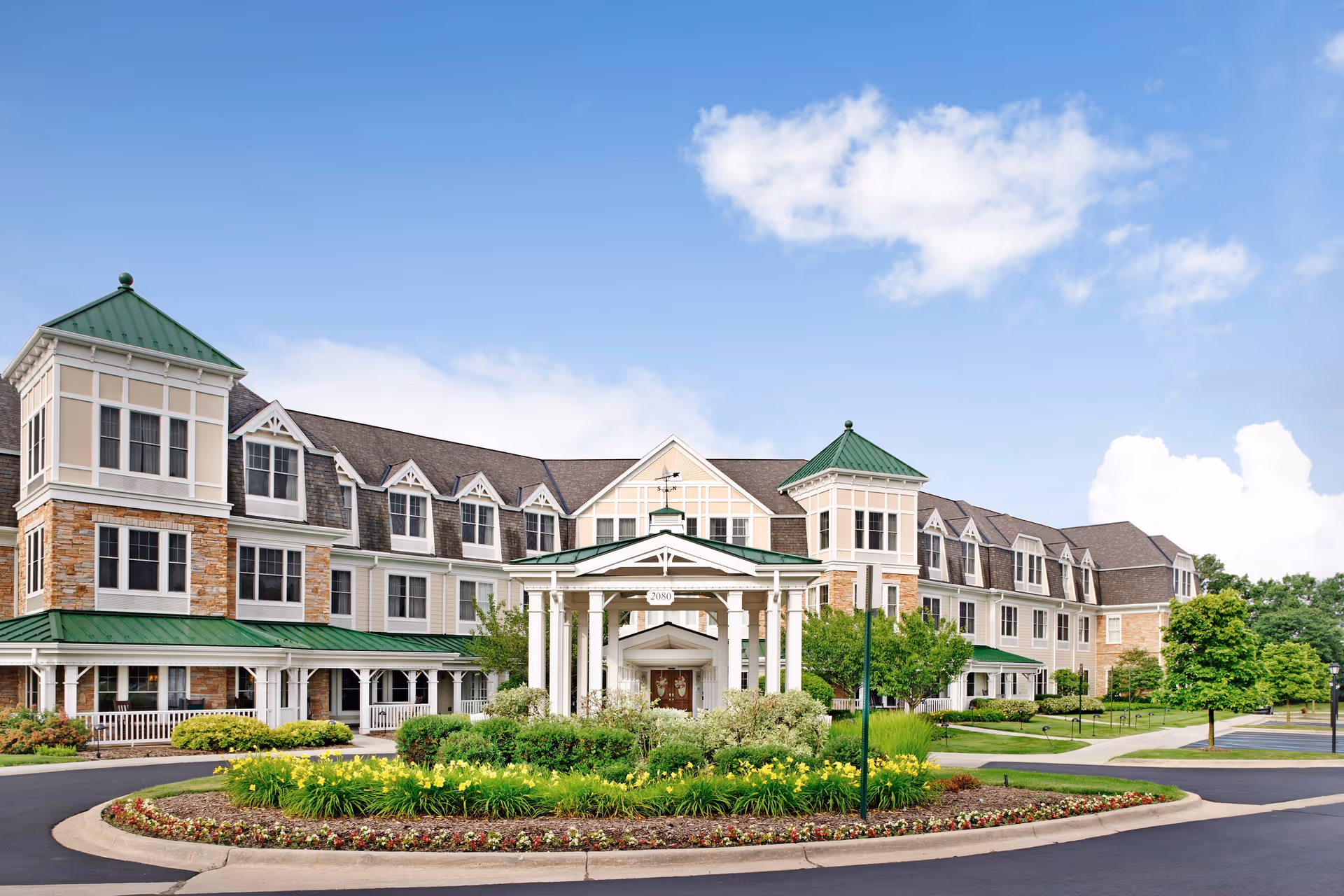 Front exterior view of The Bradford Senior Living facility featuring a large building with green roofs, multiple windows, a covered entrance, and landscaped flower beds and shrubs in the foreground under a blue sky with some clouds.