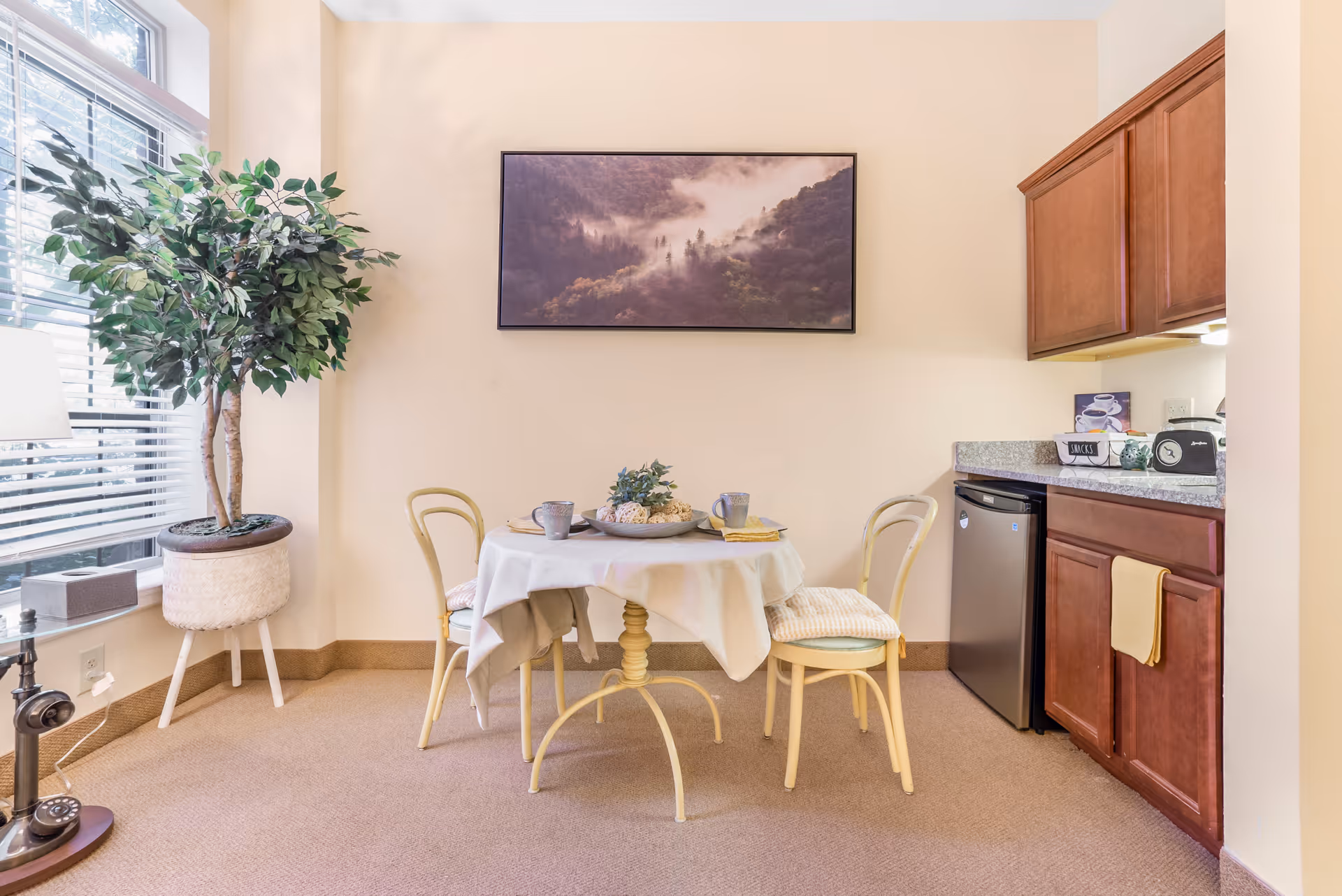 A small dining area with a round table covered with a white tablecloth, set with two cups and a decorative centerpiece. Two yellow chairs with cushions are placed around the table. To the right, there is a kitchenette with wooden cabinets, a small refrigerator, and a countertop with a toaster and other small items. A large potted plant is near a window with blinds on the left side, and a framed landscape photograph hangs on the wall above the table.