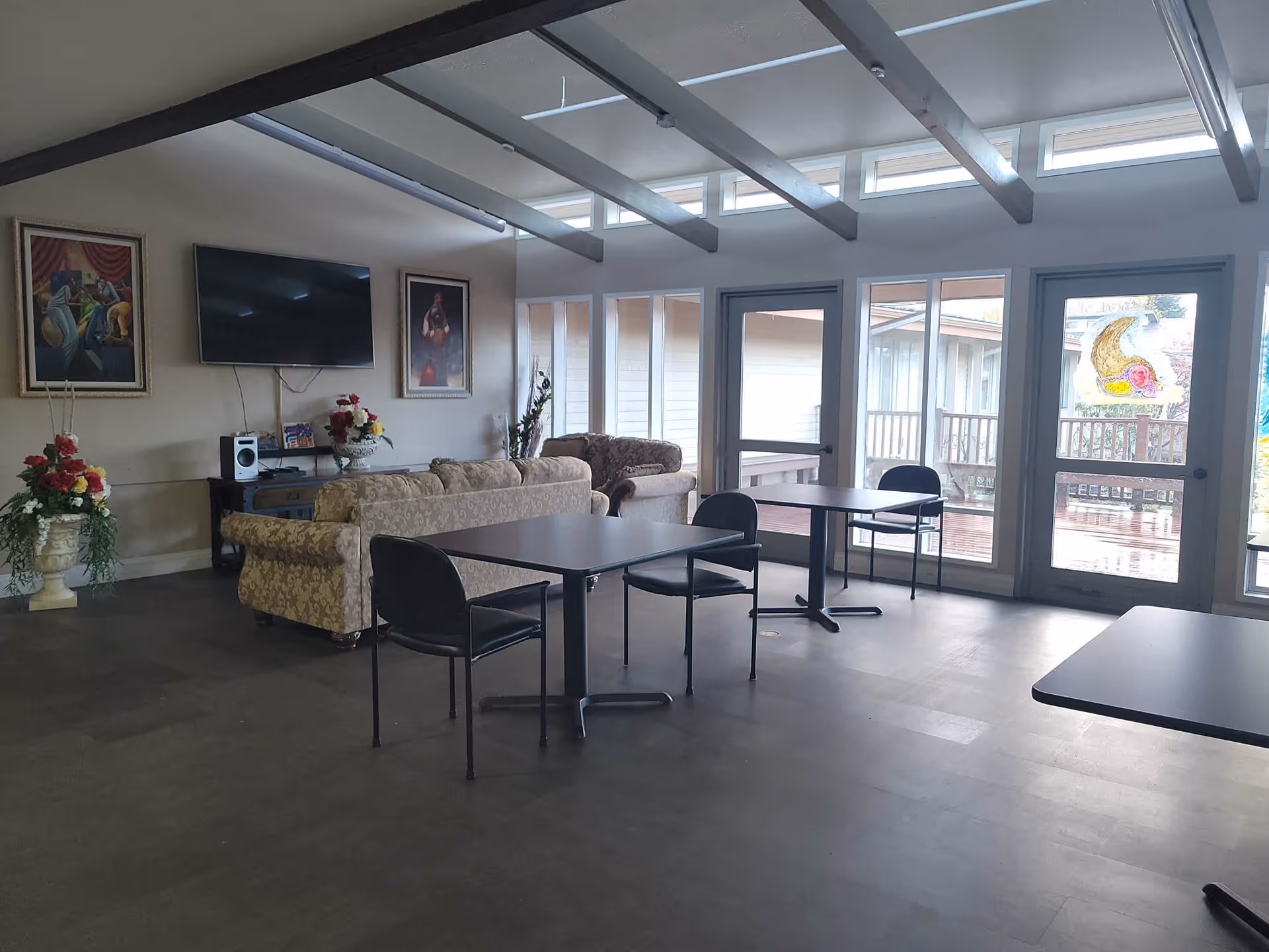 A spacious common area with several tables and chairs, two patterned sofas facing a wall-mounted flat screen TV, two framed paintings on the wall, and large windows and glass doors letting in natural light. There are decorative flower arrangements on either side of the room and visible wooden beams on the ceiling.