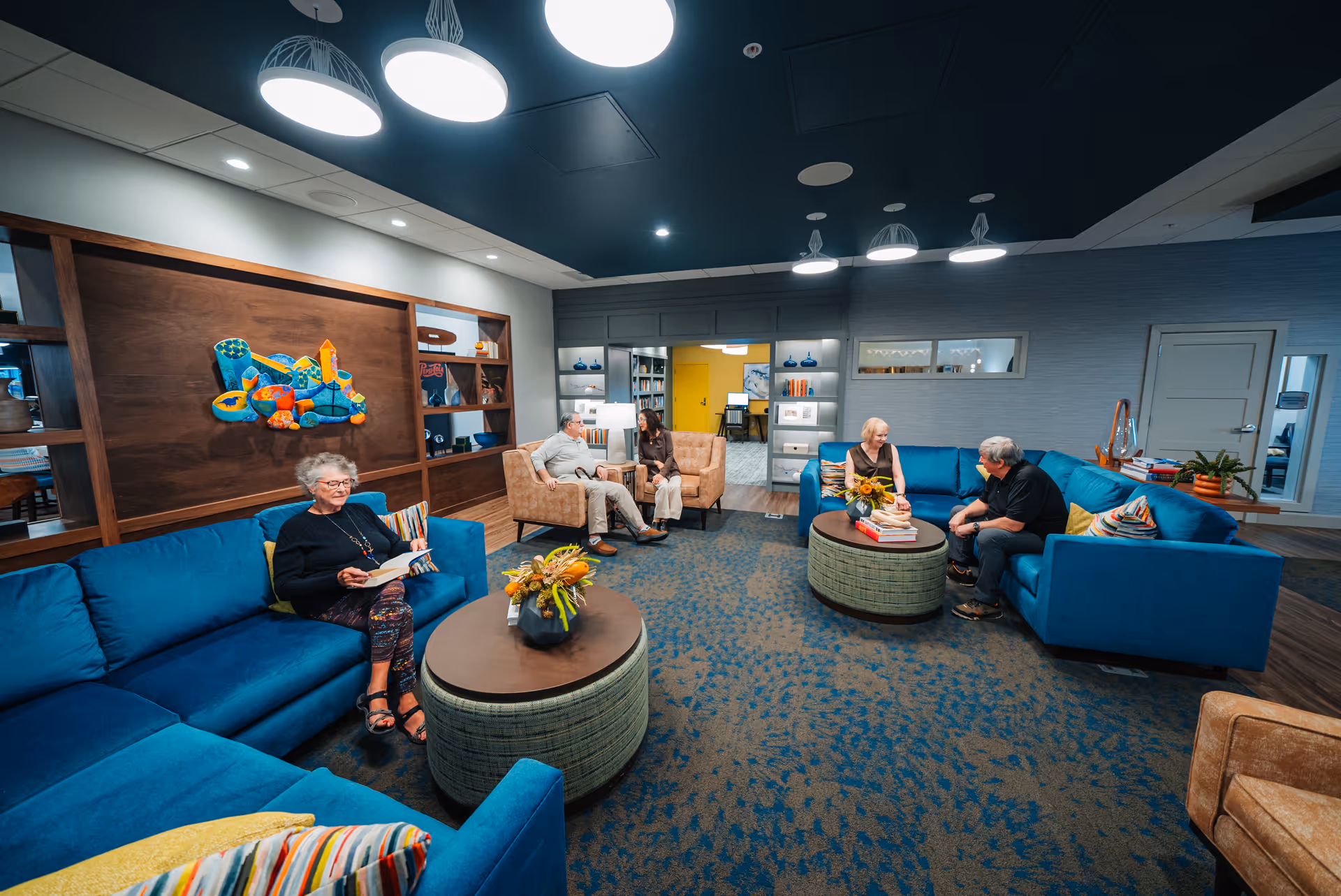 A brightly lit senior living facility lounge area with blue sofas and beige armchairs. Four elderly people are seated, two on blue sofas and two on beige armchairs, engaged in conversation or reading. The room features modern lighting fixtures, a patterned carpet, a wooden wall with colorful abstract art, and shelves with decorative items.