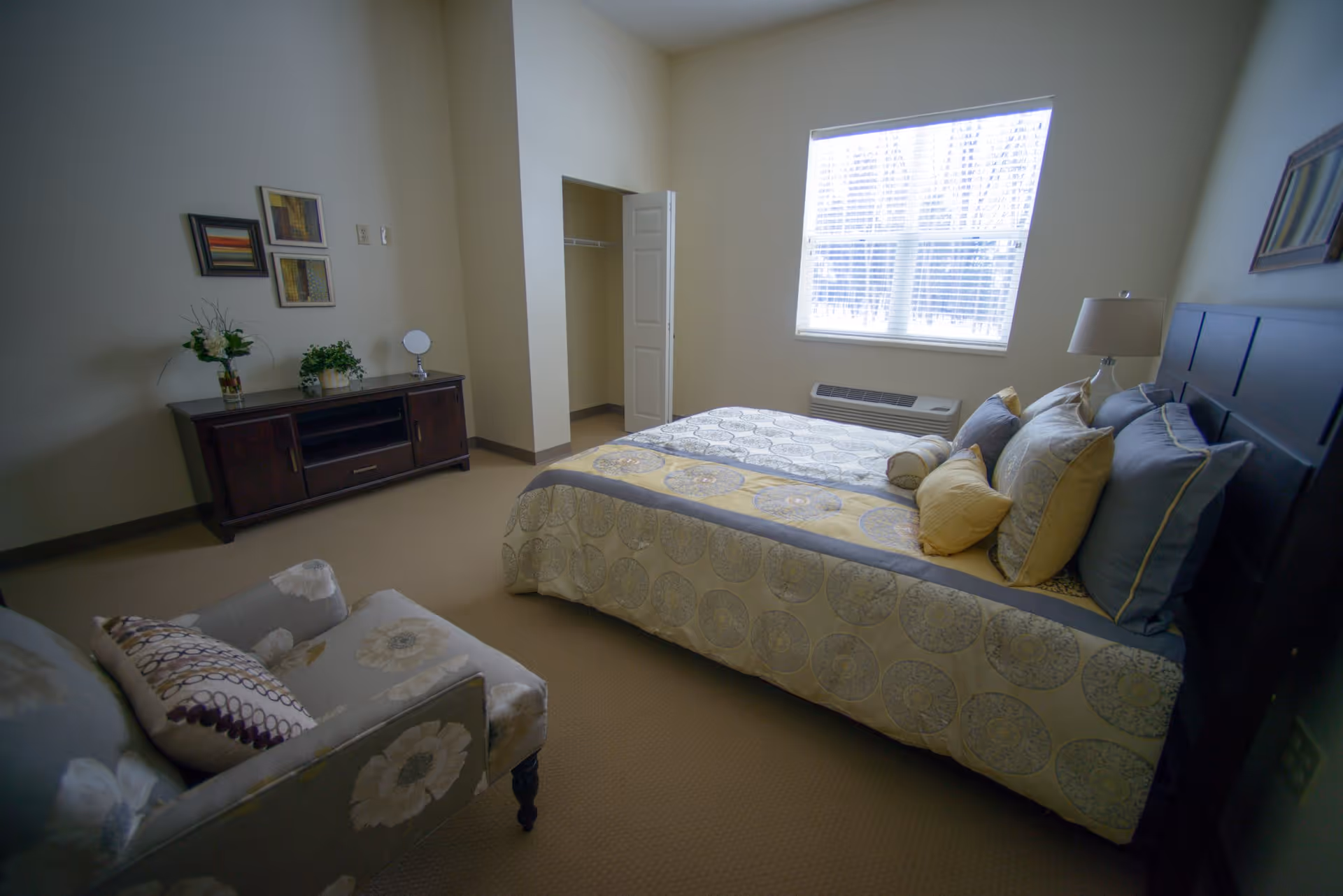 A bedroom with a bed covered in patterned bedding and multiple pillows, a floral armchair with a decorative pillow, a wooden dresser with plants and a small mirror, three framed pictures on the wall, a window with blinds letting in natural light, and an open closet door.