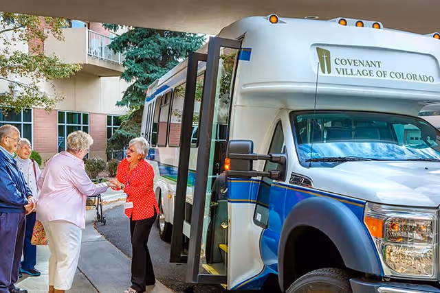 A group of elderly people standing outside near a shuttle bus labeled 'Covenant Village of Colorado.' One woman in a red polka dot blouse is greeting another woman in a light pink jacket, while two other elderly individuals stand nearby. The scene is set in front of a building with trees and bushes around.