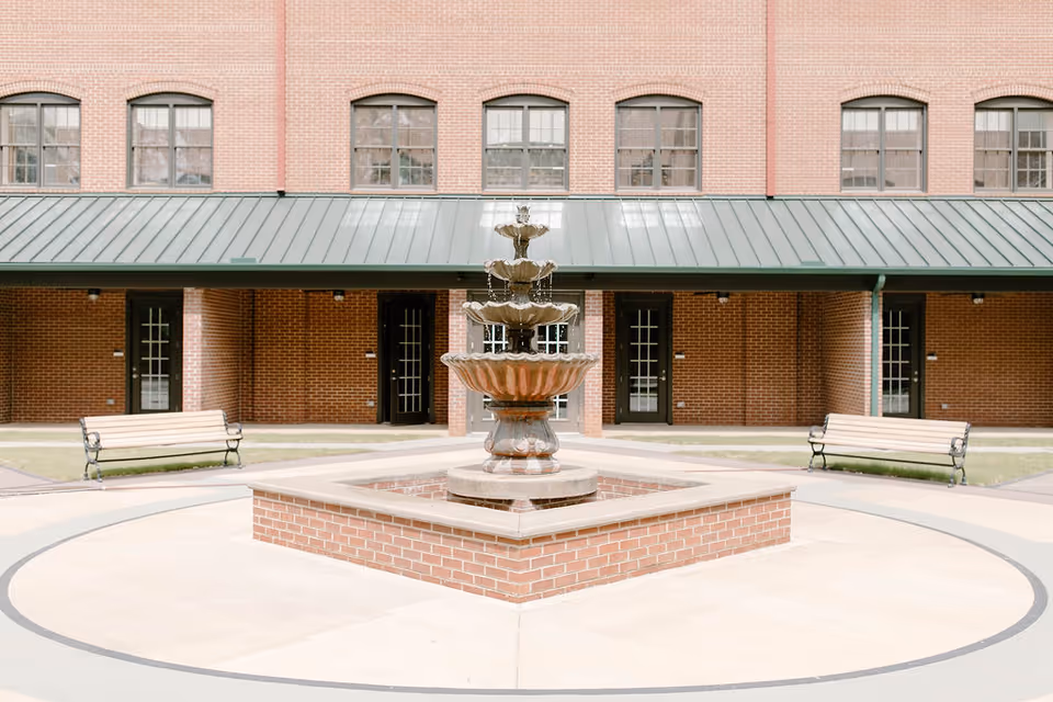 Outdoor courtyard area with a three-tiered water fountain in the center, surrounded by a circular paved walkway. There are two wooden benches with metal armrests on either side of the fountain. The background features a brick building with multiple windows and doors under a green metal awning.