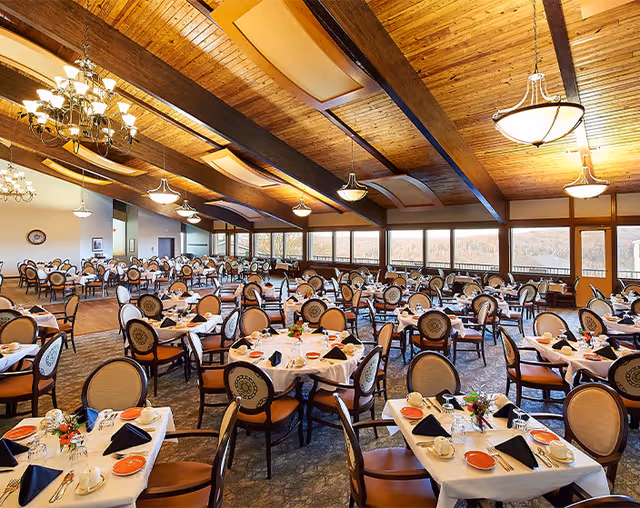 Large dining room with many round tables neatly set beneath a wooden beamed ceiling and a wall of windows.