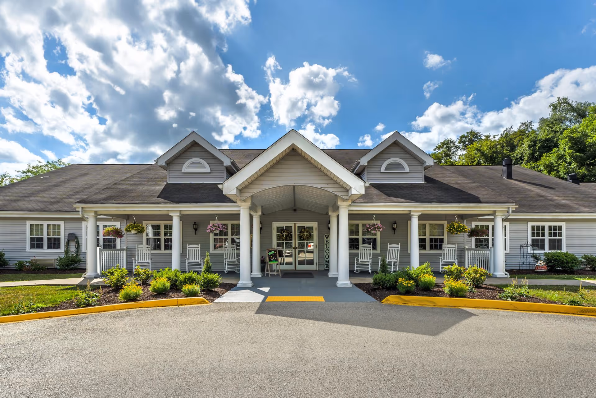 Front entrance of a single-story senior living building with a covered porte-cochère, white columns, rocking chairs, and hanging flower baskets.