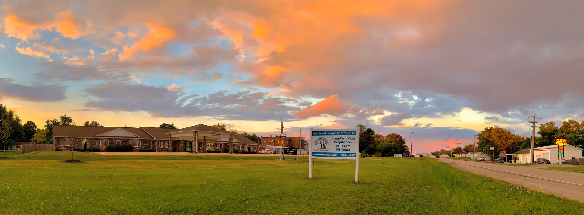 Panoramic exterior view of The Oaks Manor assisted living building set behind a wide grassy lawn with a sign and a road under a colorful sunset sky.
