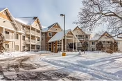 Exterior view of a multi-story senior living facility building with snow-covered ground and trees, featuring balconies and a covered entrance under a clear sky.