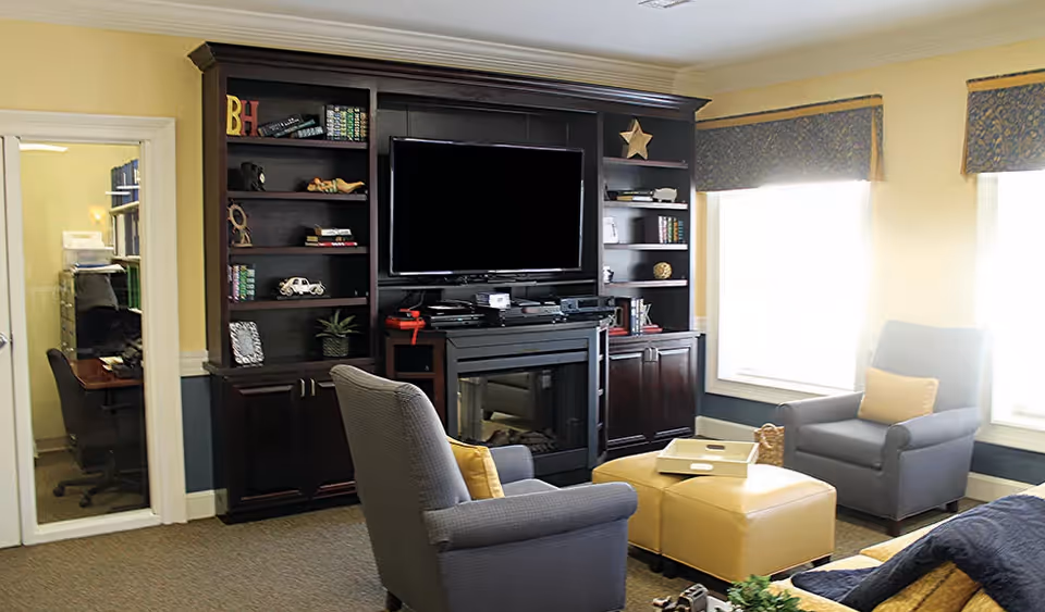 A cozy living room with two gray armchairs each with a yellow pillow, a yellow ottoman with a white tray on top, and a dark wooden entertainment center featuring a flat-screen TV and shelves with decorative items. Two large windows with patterned valances allow natural light to fill the room. A door to an office with a desk and chair is visible on the left.