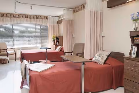 Shared senior living bedroom with two beds, red linens, curtains, and a window.
