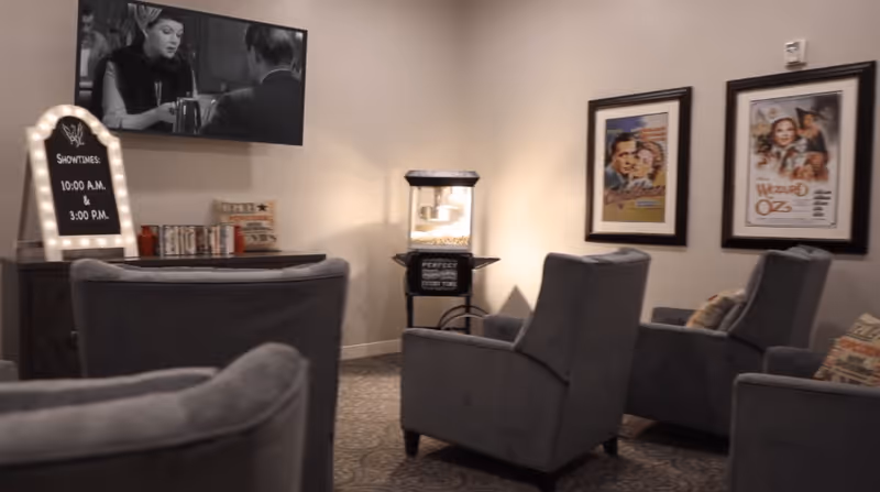 A cozy movie viewing room with several gray upholstered armchairs arranged facing a wall-mounted television playing a black-and-white film. A popcorn machine is positioned against the wall, and two framed vintage movie posters hang on the wall. A small sign with showtimes is displayed on a cabinet beneath the television.