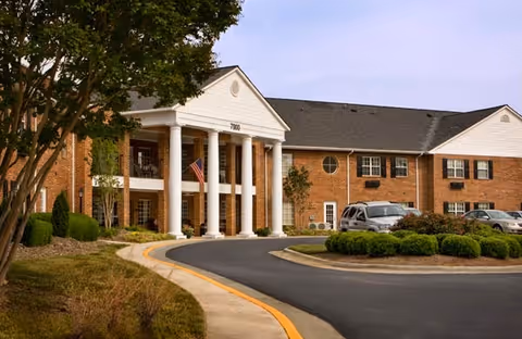 Exterior view of a senior living facility named Abbotswood at Stonehenge, featuring a brick building with white columns at the entrance, an American flag, several parked cars, and landscaped greenery along a curved driveway.