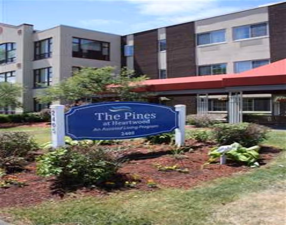 Exterior view of The Pines at Heartwood assisted living community building with a blue sign in front surrounded by landscaped bushes and grass.