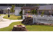 Outdoor area of a senior living facility featuring a paved walkway, a stone fire pit, a white picket fence, a pergola with chairs underneath, green grass, and a streetlamp.