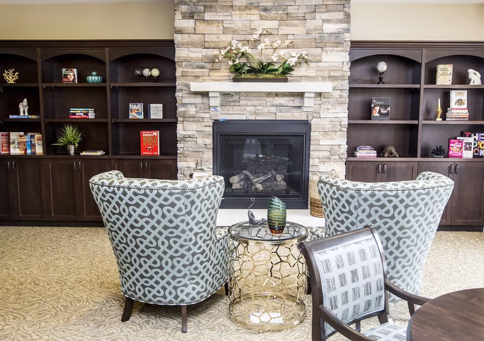 A cozy sitting area in a senior living facility featuring two patterned armchairs facing a stone fireplace with a mantel decorated with a floral arrangement. Behind the fireplace are dark wooden built-in bookshelves filled with books and decorative items. A small round glass table with a decorative piece is placed between the chairs, and part of a wooden chair and table are visible in the foreground.