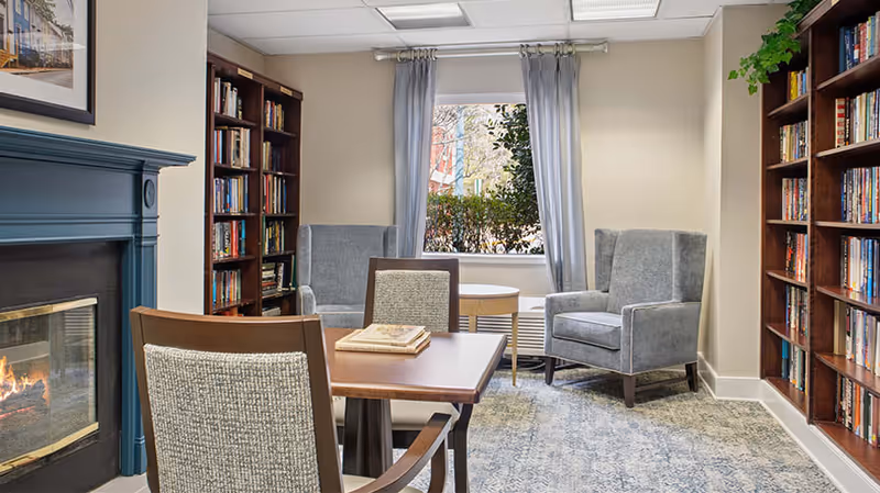 A cozy senior living community common area with a lit fireplace on the left, two tall bookshelves filled with books on either side, a wooden table with two chairs in the foreground, and two gray armchairs near a window with light gray curtains. A small round table is placed between the armchairs.
