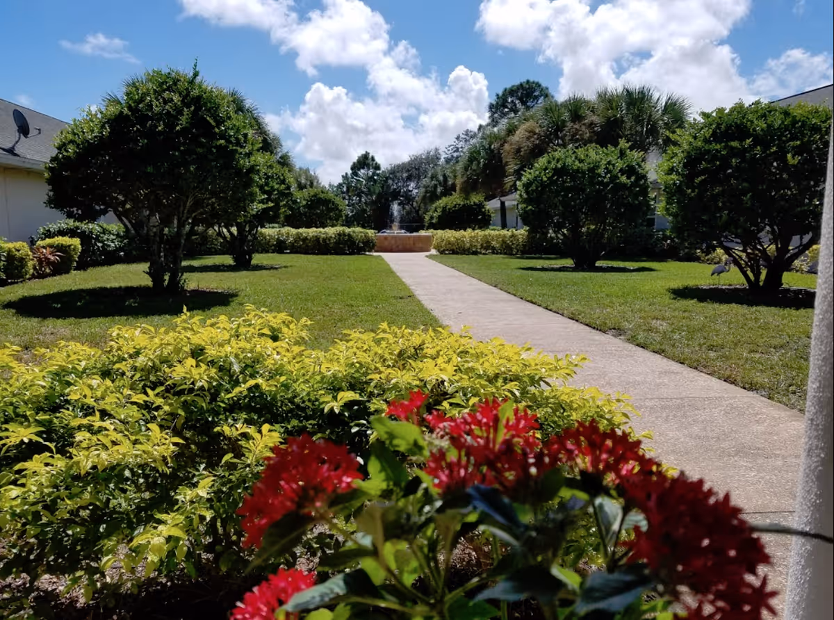 A sunny outdoor garden area with a concrete pathway leading to a water fountain in the distance. The garden is surrounded by neatly trimmed bushes and trees, with vibrant red flowers in the foreground. The sky is blue with scattered white clouds.
