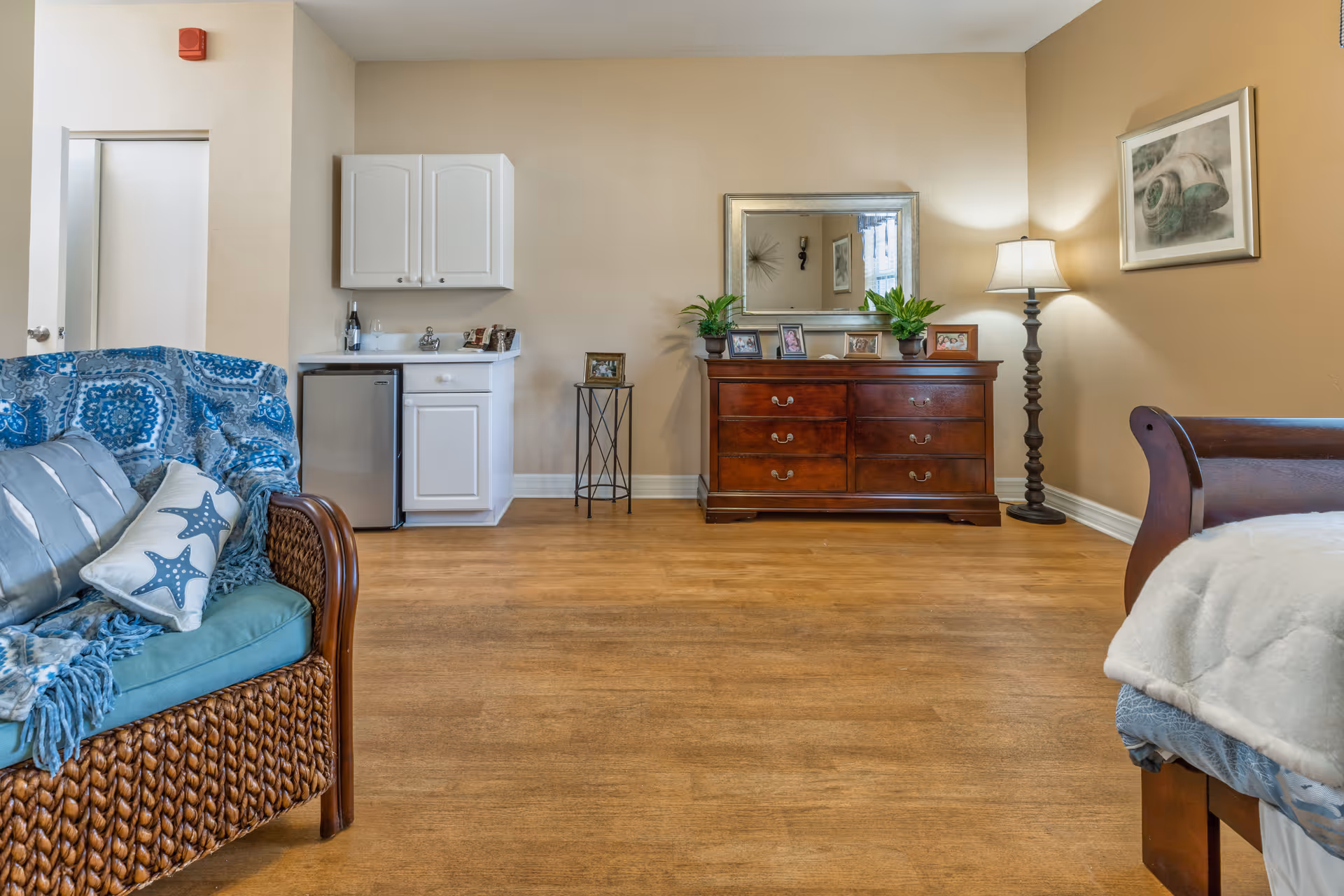 Furnished senior living suite with a wicker sofa, wooden dresser and mirror, floor lamp, small kitchenette and a bed partially visible on the right.