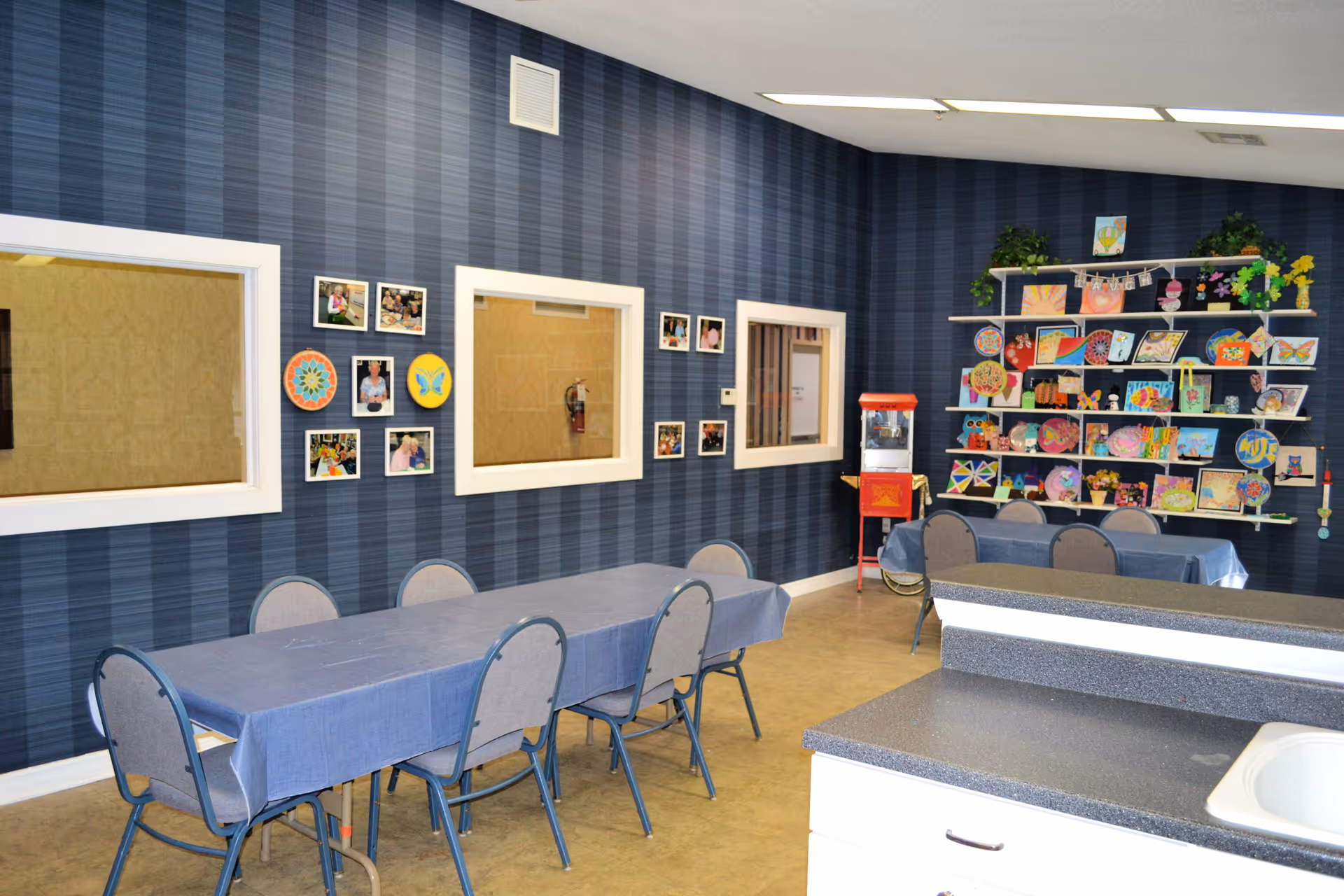 Community activity/dining room with tables and chairs, craft display shelves, and framed windows on a blue-striped wall.