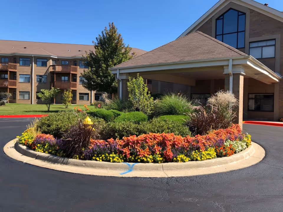 Entrance area of Bridgeway of Bensenville Supportive Living facility featuring a circular flower bed with colorful flowers and shrubs, a covered drop-off area, and a multi-story brick building in the background under a clear blue sky.