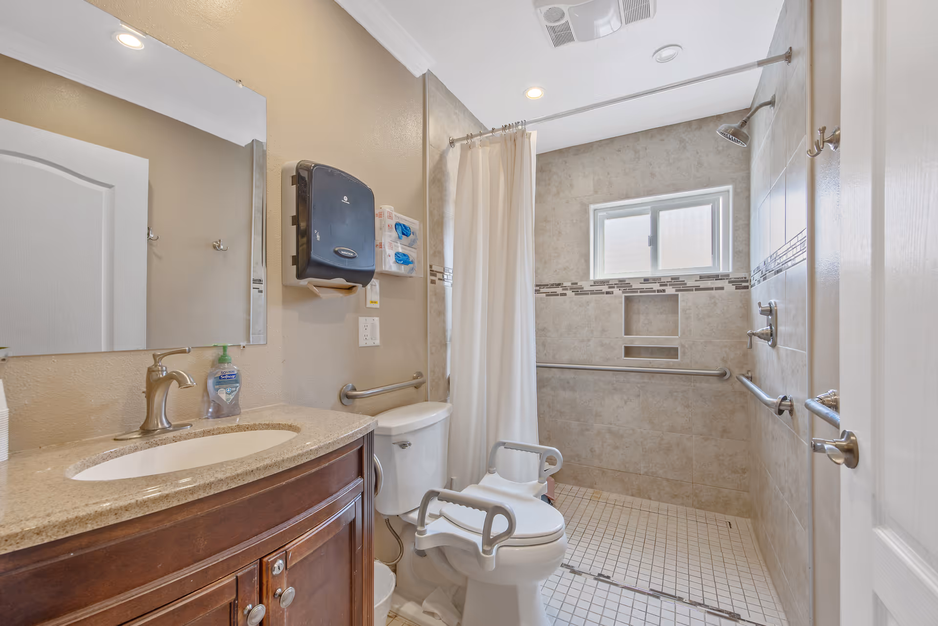 A bathroom with beige walls and tiled shower area featuring grab bars and a handheld showerhead. There is a white toilet with arm supports, a wooden vanity with a sink and faucet, a large mirror above the sink, and a paper towel dispenser mounted on the wall. The shower has a curtain and a small window for natural light.
