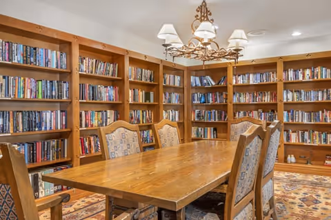 A cozy library room with wooden bookshelves filled with books lining the walls. In the center, there is a large wooden table surrounded by six upholstered chairs with patterned fabric. A decorative chandelier with lampshades hangs above the table, and a patterned rug covers the floor.
