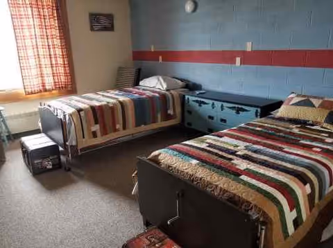 A simple bedroom with two single beds, each covered with a colorful patchwork quilt. The room has a window with checkered curtains allowing natural light to enter. There is a blue chest of drawers between the beds and a small suitcase on the floor near each bed. The walls are painted with a blue and red stripe design.
