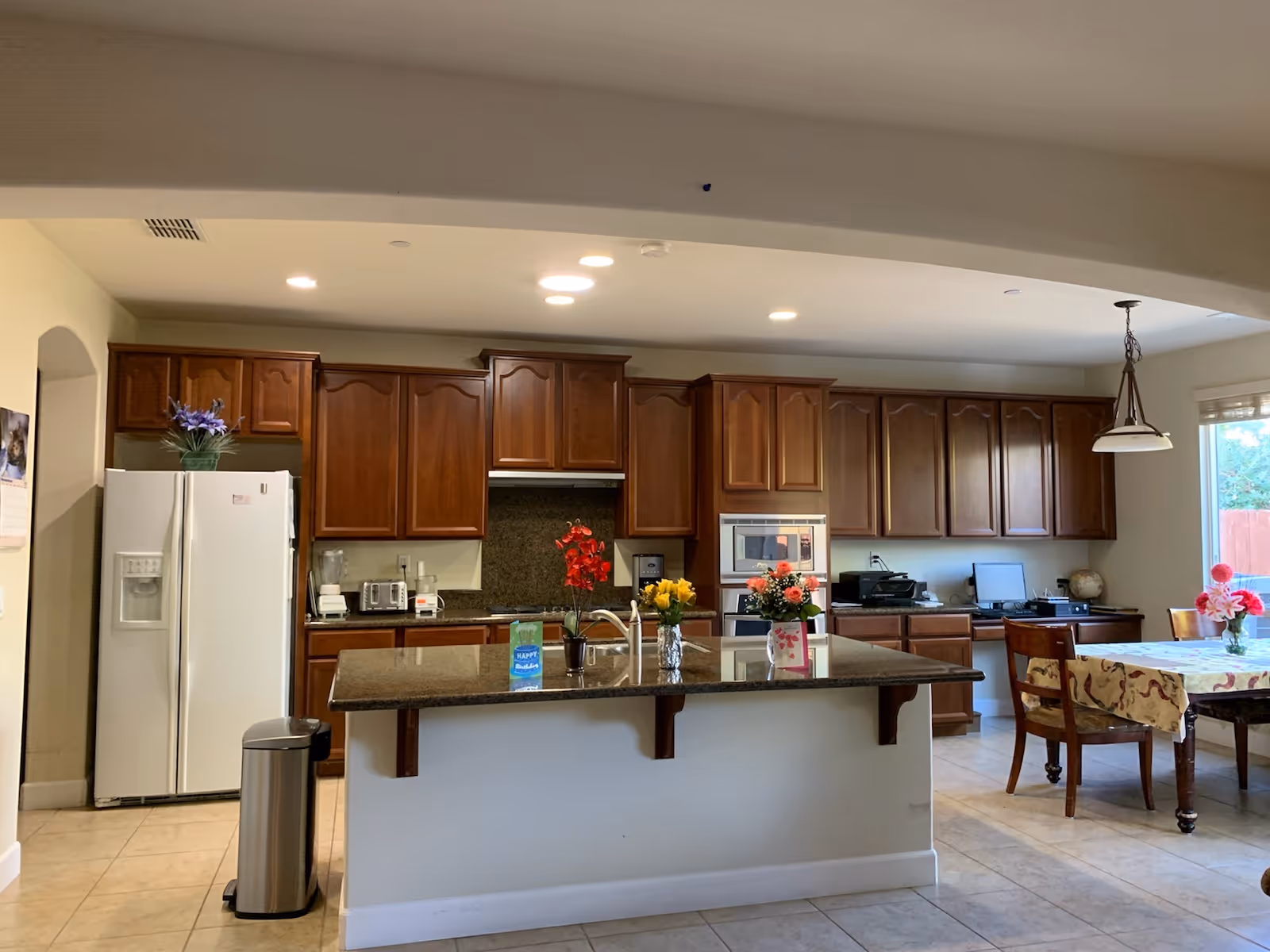 A spacious kitchen with wooden cabinets, a white refrigerator, a granite countertop island with a sink, and various flowers in vases placed on the island. There is a dining table with chairs and a hanging light fixture near a window with a view of a fenced backyard.