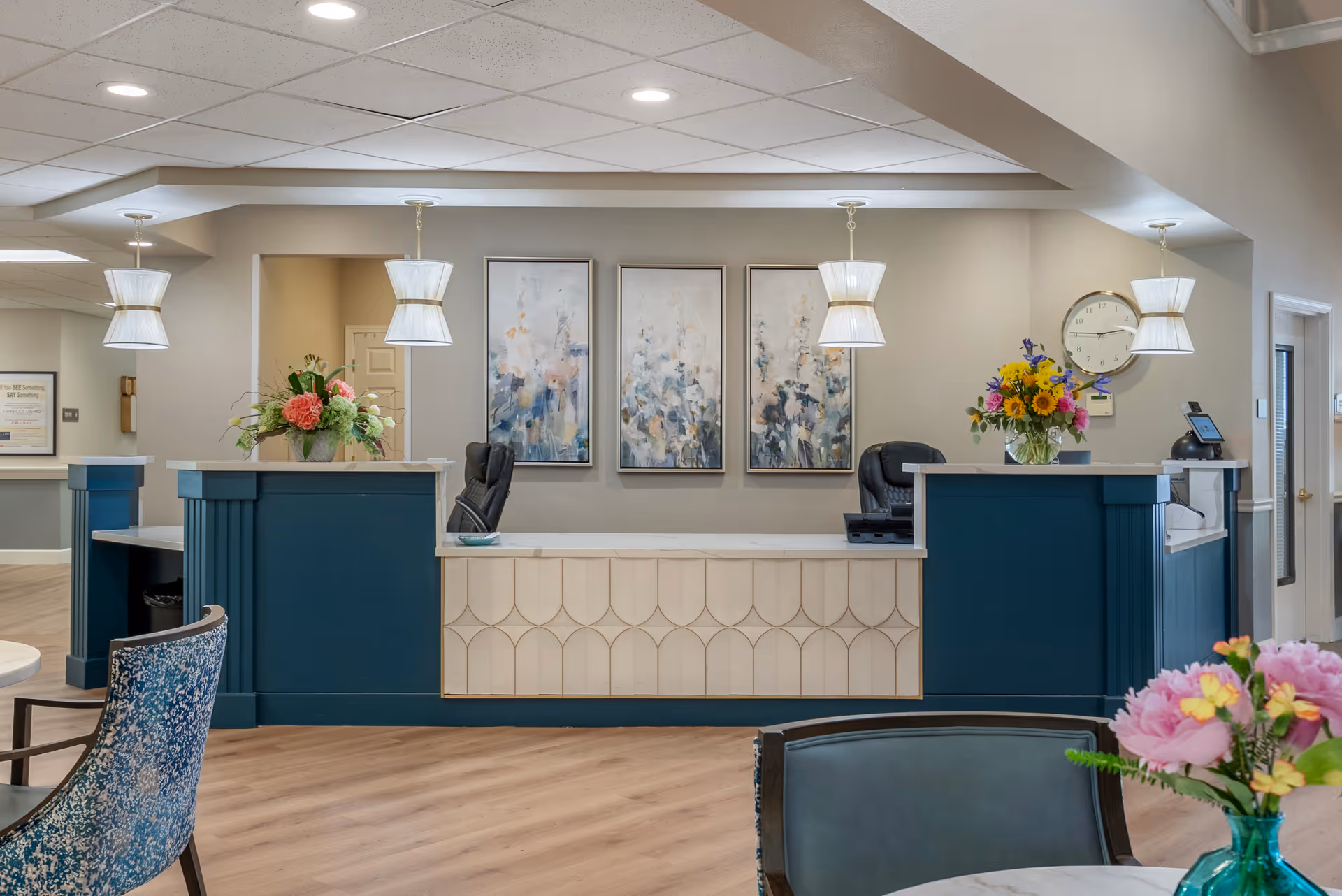 Reception desk area in a senior living facility lobby with teal counters, pendant lights, floral arrangements, and framed artwork on the wall.