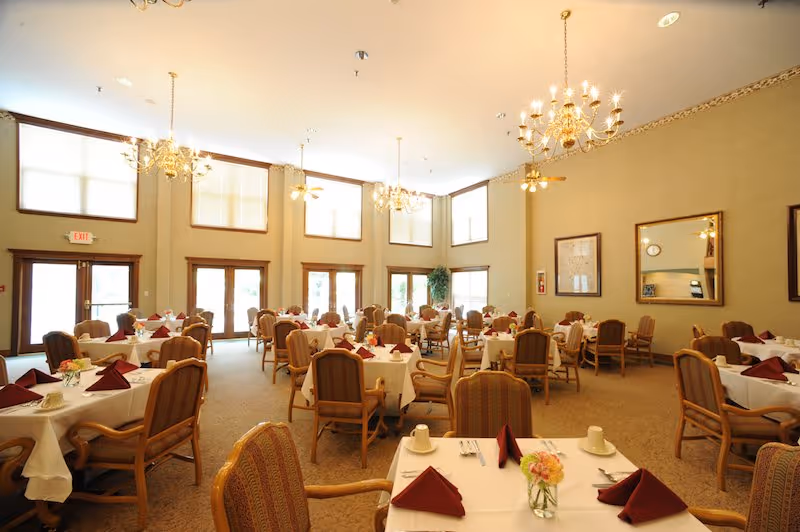 A spacious dining room with multiple round tables covered with white tablecloths, each set with maroon folded napkins, cups, and silverware. The room features high ceilings with large windows letting in natural light, elegant chandeliers, and comfortable cushioned chairs. There are mirrors and framed pictures on the beige walls.