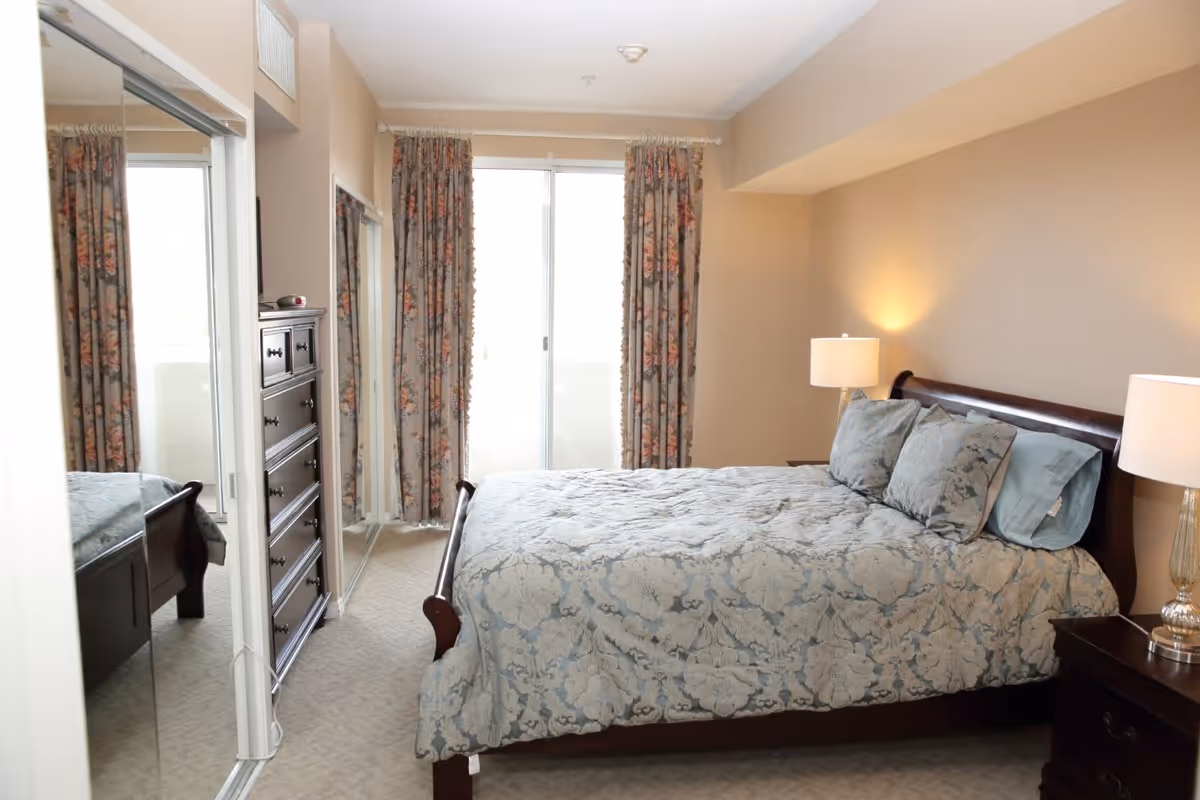 A bedroom with a large bed covered in a blue and beige floral patterned comforter and matching pillows. There are two bedside tables with lamps on each side of the bed. A tall dark wooden dresser is positioned against the wall near a sliding glass door with floral curtains. A large mirror is mounted on the wall next to the dresser, reflecting part of the bed.