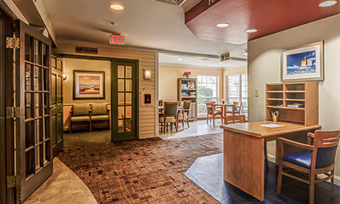 Interior view of a senior living facility showing a reception desk with a chair, a seating area with a couch and framed artwork on the wall, and a dining area with tables and chairs near large windows letting in natural light.