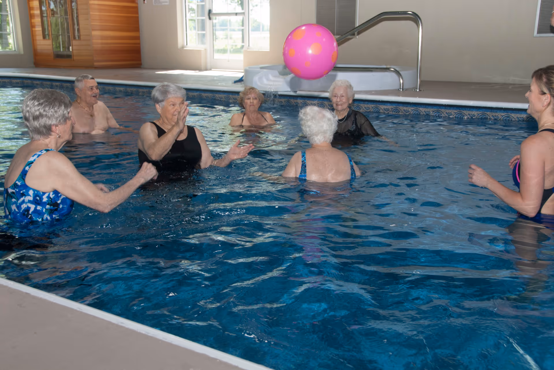 A group of elderly people and a younger woman playing with a pink polka dot beach ball in an indoor swimming pool at Bethea Retirement Community.