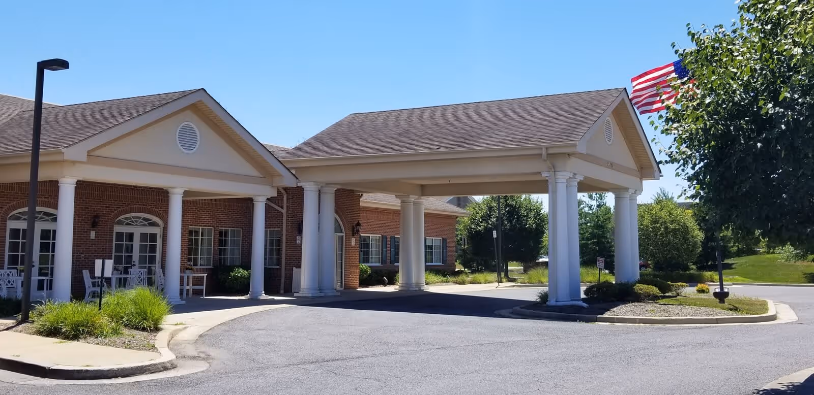 Exterior view of Meadow Park Rehabilitation And Healthcare Center showing a covered entrance with white columns, a brick building, an American flag, and surrounding greenery under a clear blue sky.